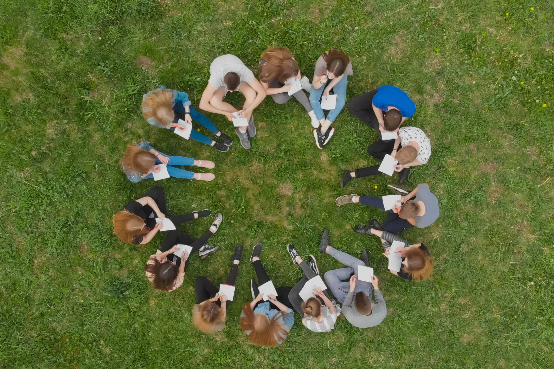Menschen sitzen im Kreis auf einer Wiese. Die Aufnahme ist aus der Vogelpersepktive aufgenommen.