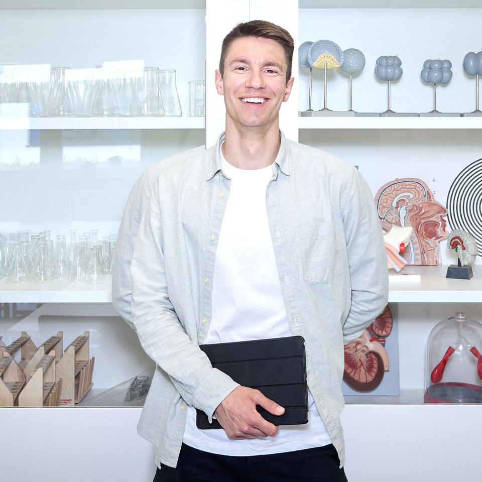 Lehrkraft Sebastian Engel steht im Biologieraum vor einer Glasvitrine und schaut in die Kamera.