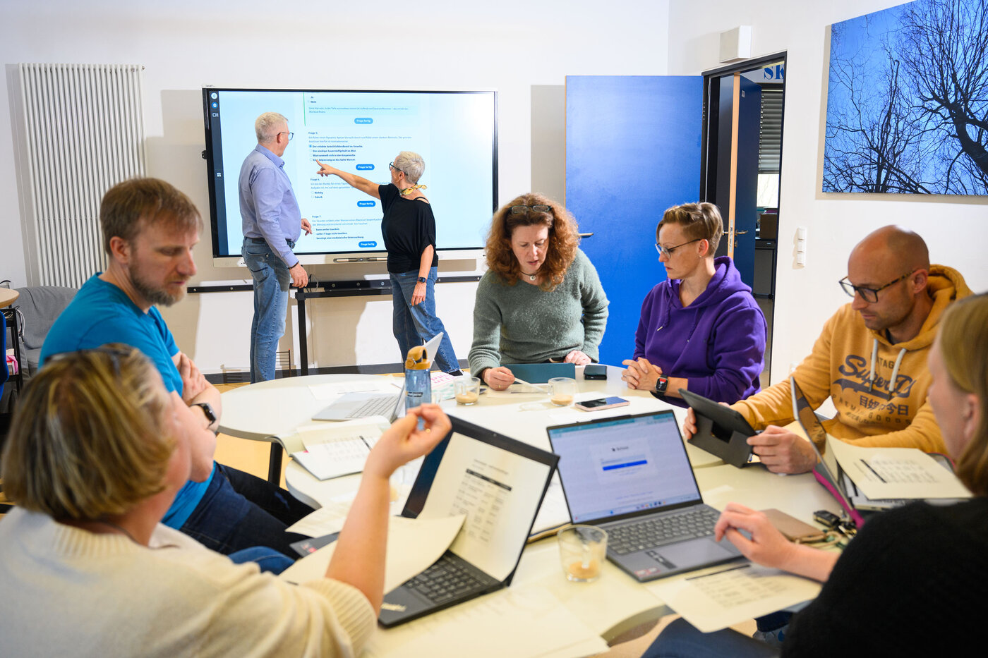 Erwachsene sitzen um einen Tisch mit Laptops. Im Hintergrund stehen zwei Erwachsene vor einem Smartboard.
