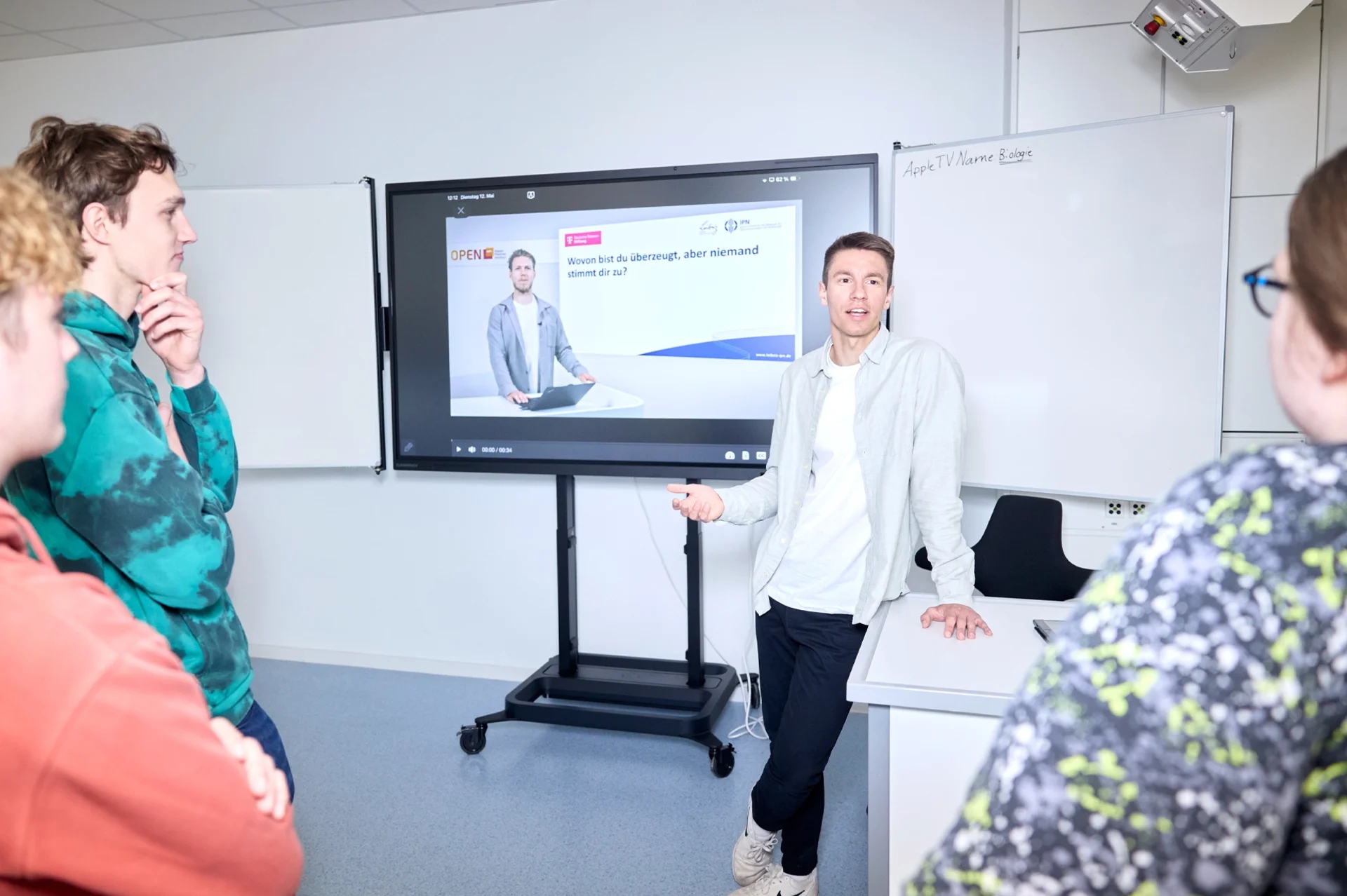 Lehrkraft Sebastian Engel spricht im Biologieraum mit älteren Schüler*innen, im Hintergrund läuft auf einem Screen ein Onlinetutorial.