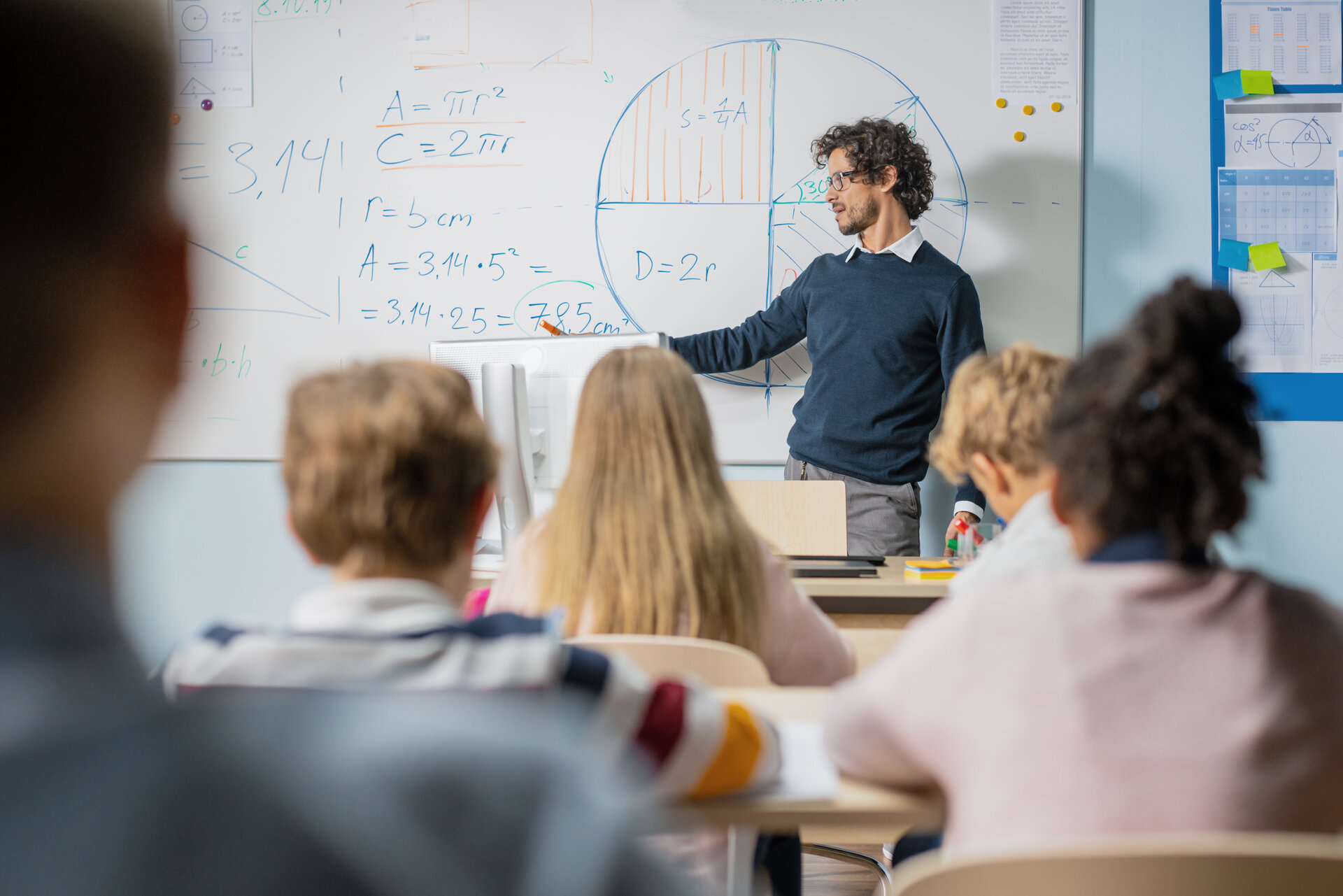 Eine Lehrkraft steht vor einer Klasse und erklärt etwas an der Tafel.