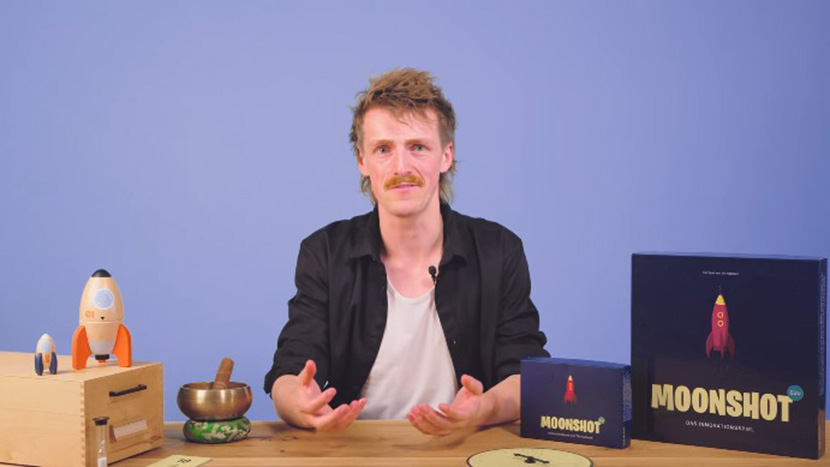 Till Hasbach sitzt an einem Tisch und hat vor sich das Spiel Moonshot EDU liegen.