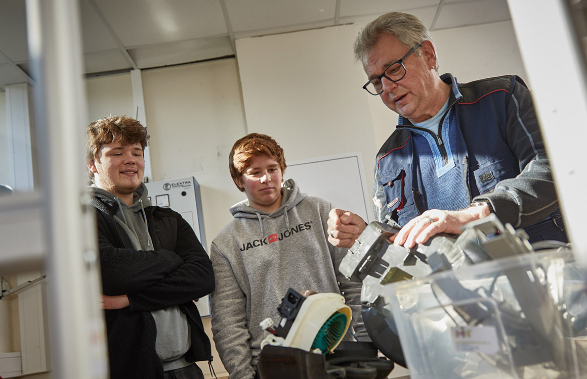 Das Bild zeigt Andreas Milchner, der mit zwei Schülern ein elektronisches Gerät repariert.