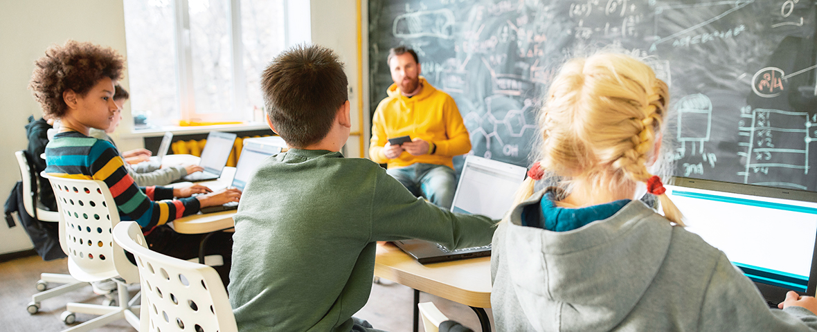 Drei Schüler schauen zu Lehrkraft, der am Pult sitzt.