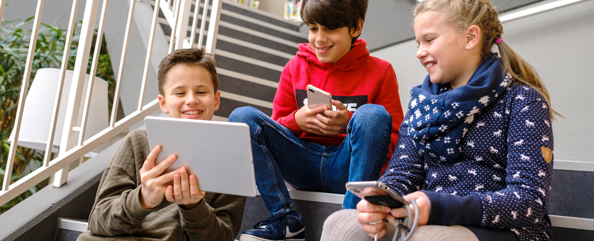 Das Bild zeigt drei Kinder, die mit Laptop und Smartphones auf einer Treppe sitzen.