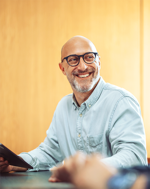 Ein Mann sitzt an einem Tisch, hat ein Tablet in der Hand und lacht.