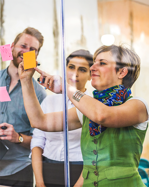 Drei Erwachsene stehen vor einer Glaswand und kleben Post-its.