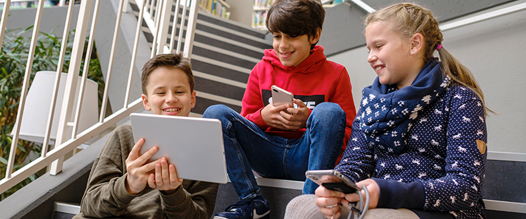 Das Bild zeigt drei Kinder, die mit Laptop und Smartphones auf einer Treppe sitzen.
