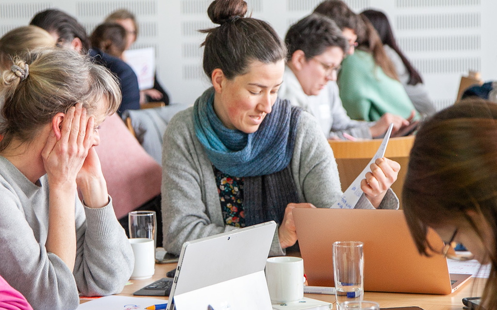 Erwachsene sitzen gemeinsam an einem Tisch und haben ihre Laptops und Tablets vor sich stehen.