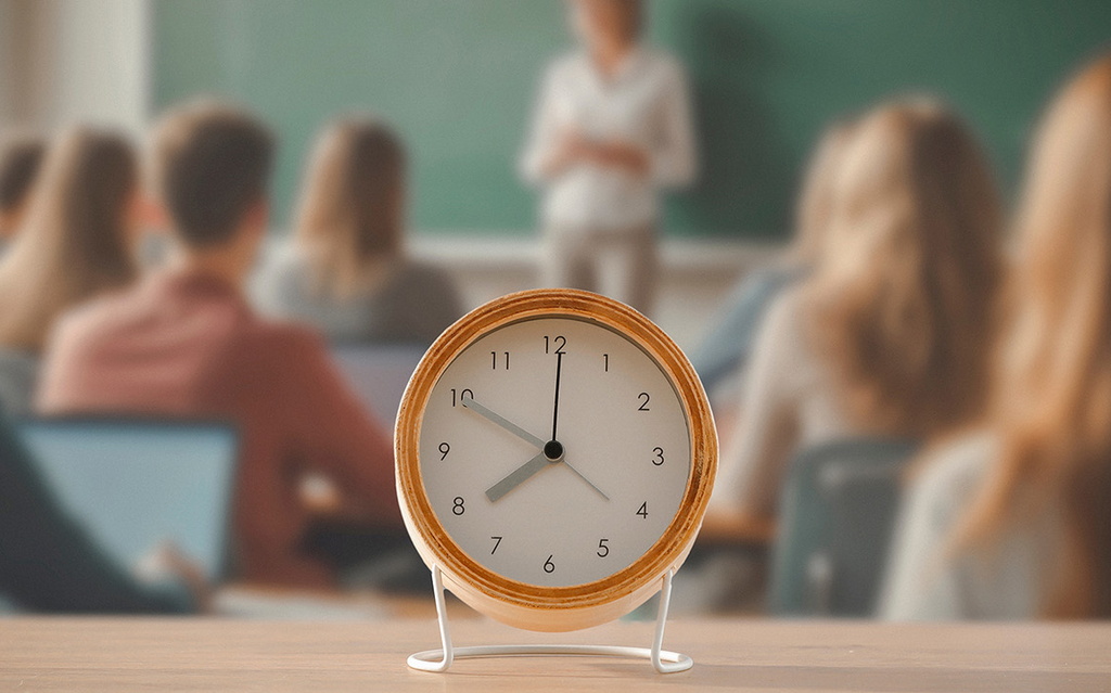 Im Vordergrund ist ein Wecker zu sehen, der auf einem Tisch steht. Dahinter ist eine Klassenraum mit Schüler:innen und einer Lehrkraft zu sehen.