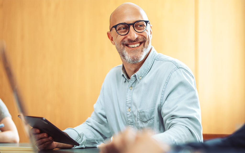 Ein Mann sitzt an einem Tisch, hat ein Tablet in der Hand und lacht.