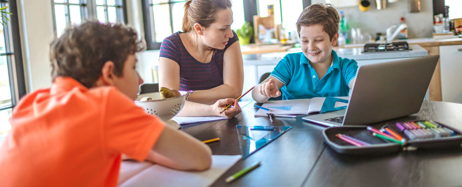 The picture shows two boys learning together with their mother at the kitchen table.
