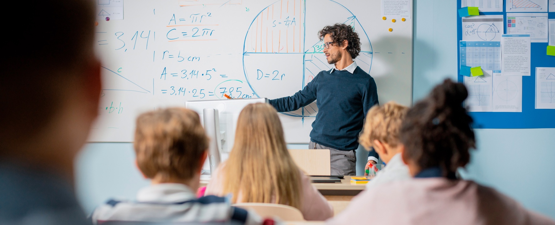 The picture shows a teacher standing in front of a class at the blackboard.