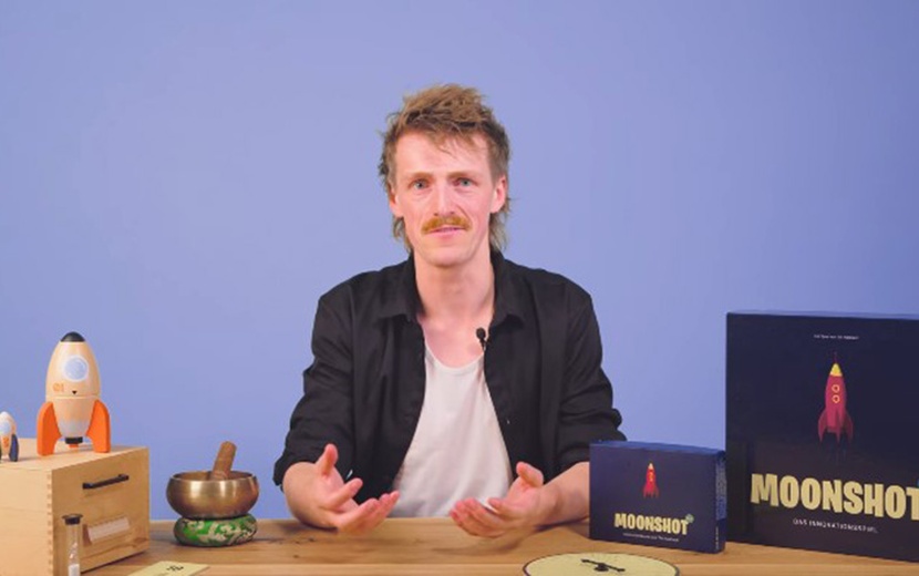 Till Hasbach sitzt an einem Schreibtisch, auf dem das Spiel "Moonshot EDU" liegt.