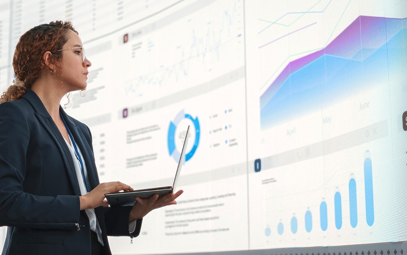 Eine Frau mit einem Laptop in der Hand steht vor einer großen Bildschirmwand.