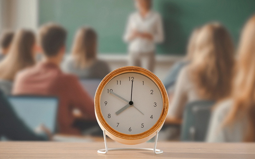 Im Vordergrund ist ein Wecker zu sehen, der auf einem Tisch steht. Dahinter ist eine Klassenraum mit Schüler:innen und einer Lehrkraft zu sehen.