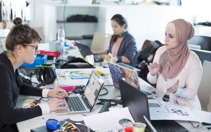 Mädchen sitzen an Arbeitstisch vor Laptops und sprechen miteinander.