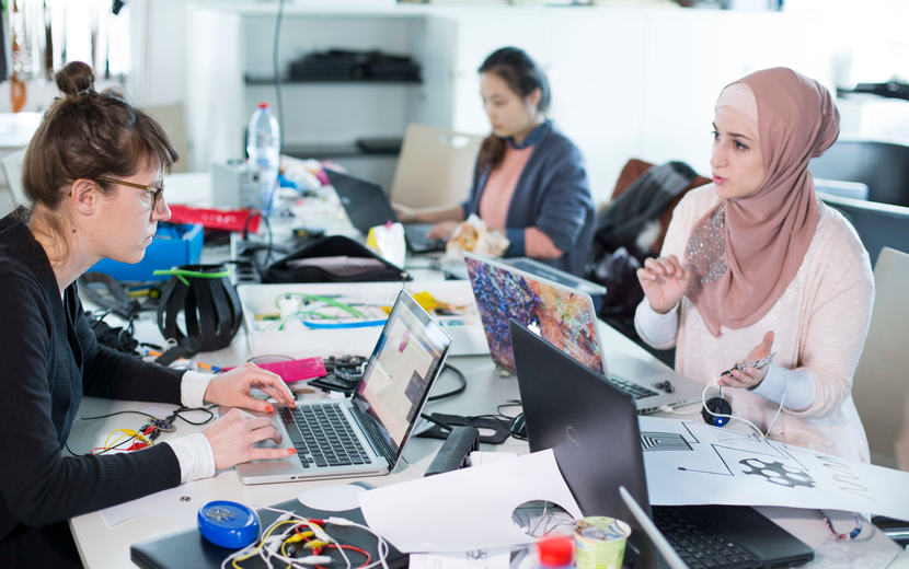 Mädchen sitzen an Arbeitstisch vor Laptops und sprechen miteinander.