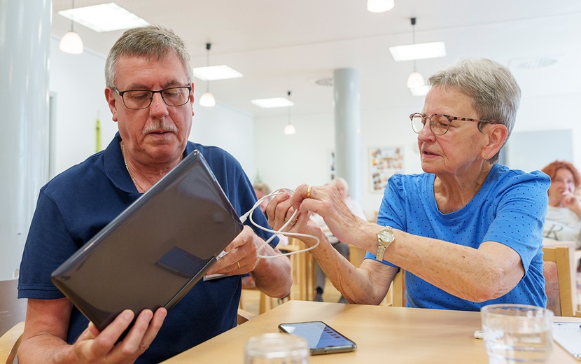 Eine ältere Dame hilft einem älteren Herren mit seinem Laptop.